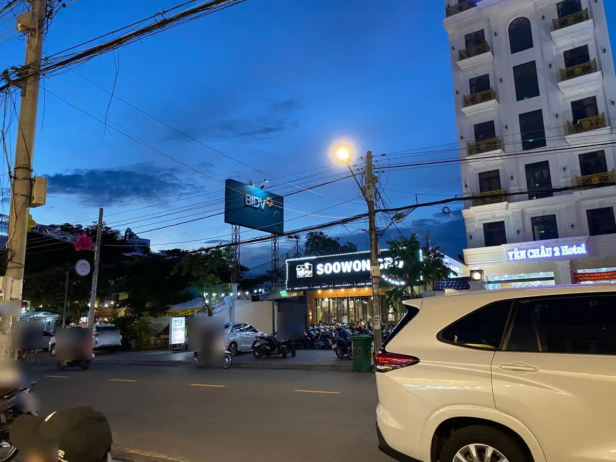 Evening street with motorbikes passing parked scooters outside shops and a hotel, under street lights and power lines