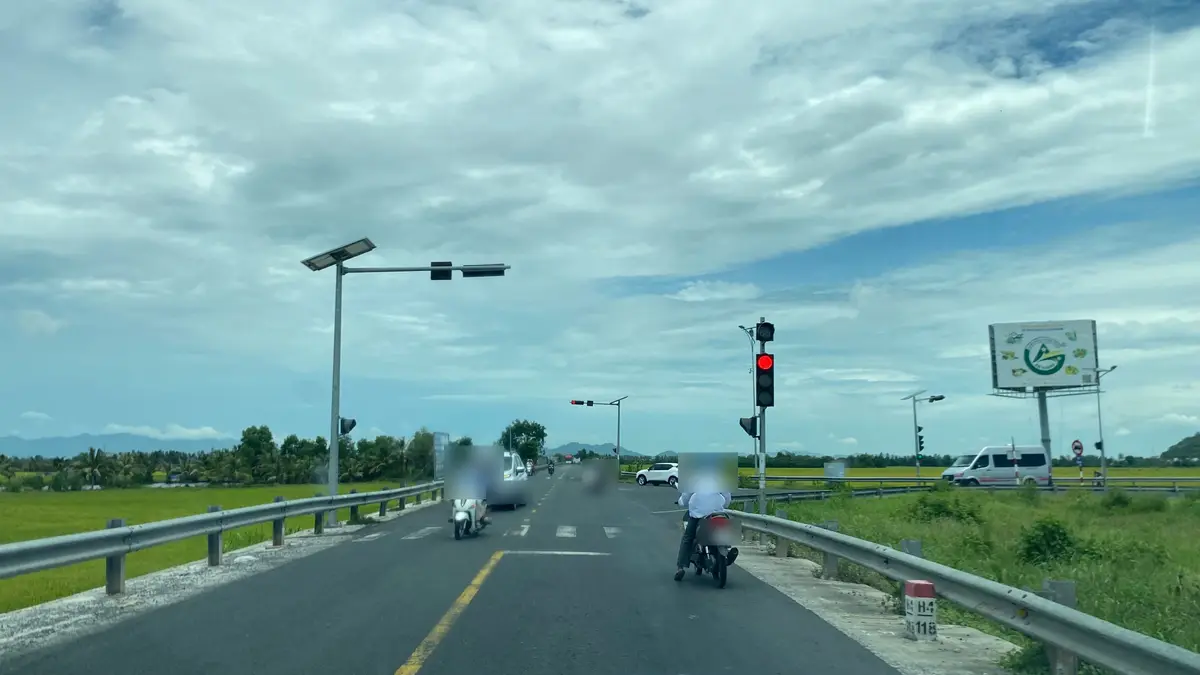 Two-lane rural road at a red traffic light with motorbikes and cars, rice fields on both sides under cloudy sky