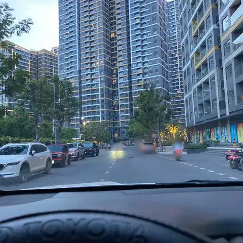 Street between high-rise apartment towers at dusk, seen from a car's driver seat, with parked cars and a passing motorbike