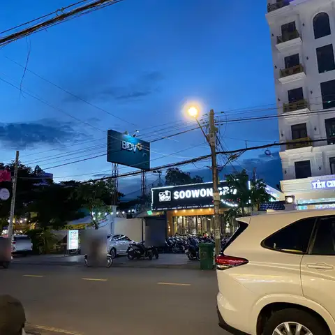 Evening street with motorbikes passing parked scooters outside shops and a hotel, under street lights and power lines
