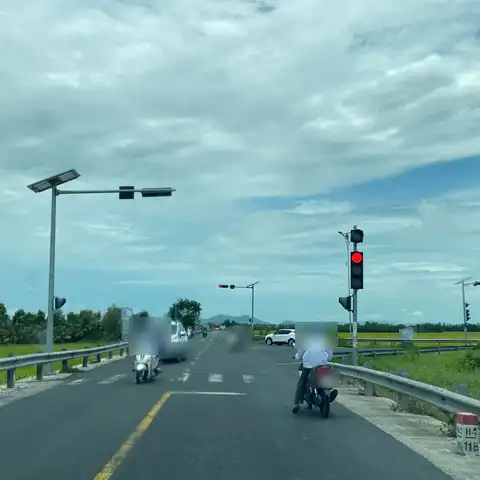 Two-lane rural road at a red traffic light with motorbikes and cars, rice fields on both sides under cloudy sky