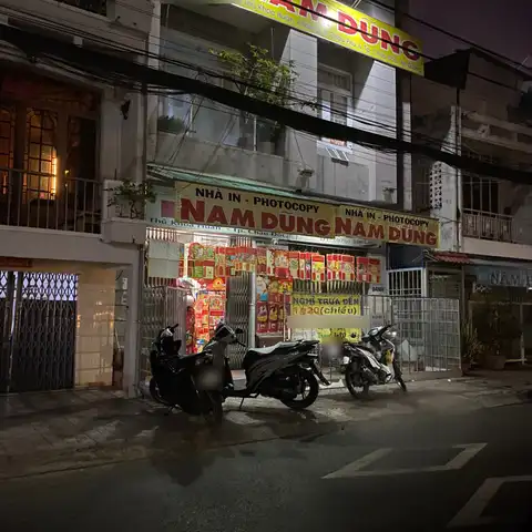 Night street view of a print and photocopy shop with large Vietnamese signs reading 'Nhà in - Photocopy Nam Dũng' and parked motorbikes